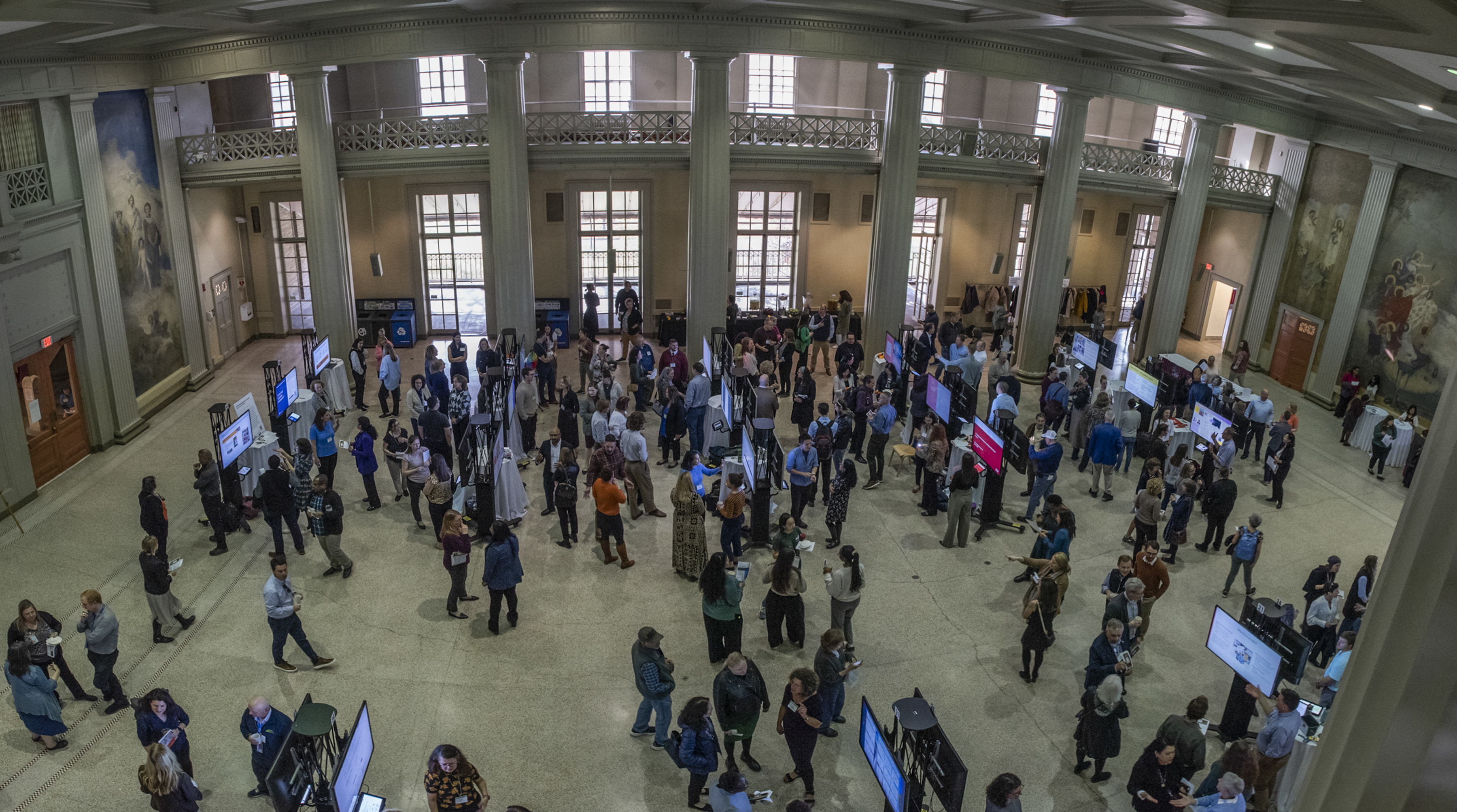 Poster session attendees are engaged in conversation around digital displays in Morss Hall, a spacious room with tall windows, columns, and murals.